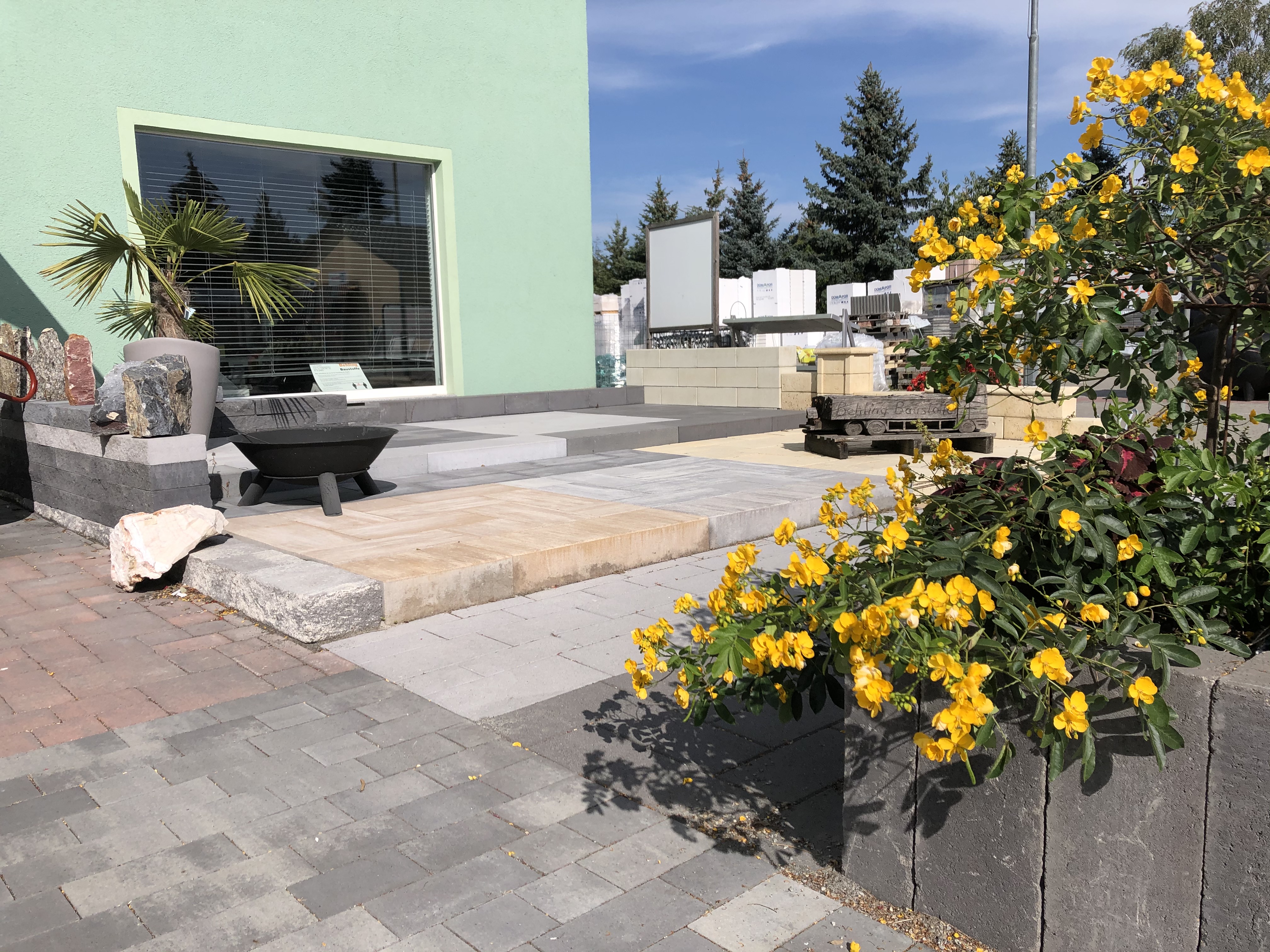 Moderne Gartengestaltung mit Steinplatten, einer Feuerschale und blühenden gelben Blumen vor einem Gebäude